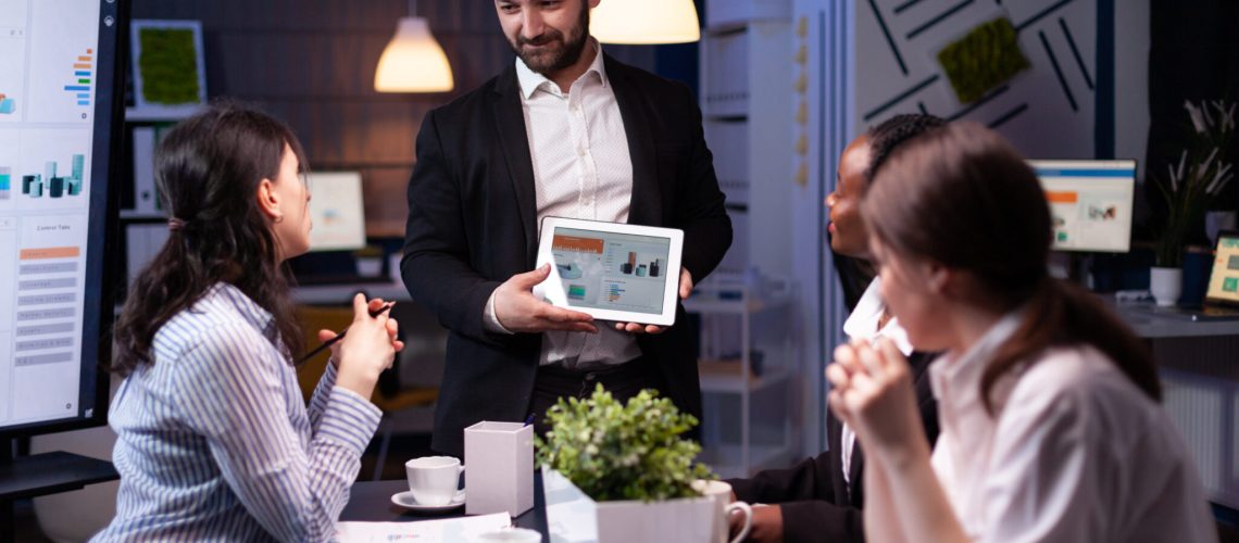 Entrepreneur businessman showing company strategy using tablet for corporate presentation working overtime in meeting office room late at night. Diverse multi-ethnic teamwork brainstorming ideas.